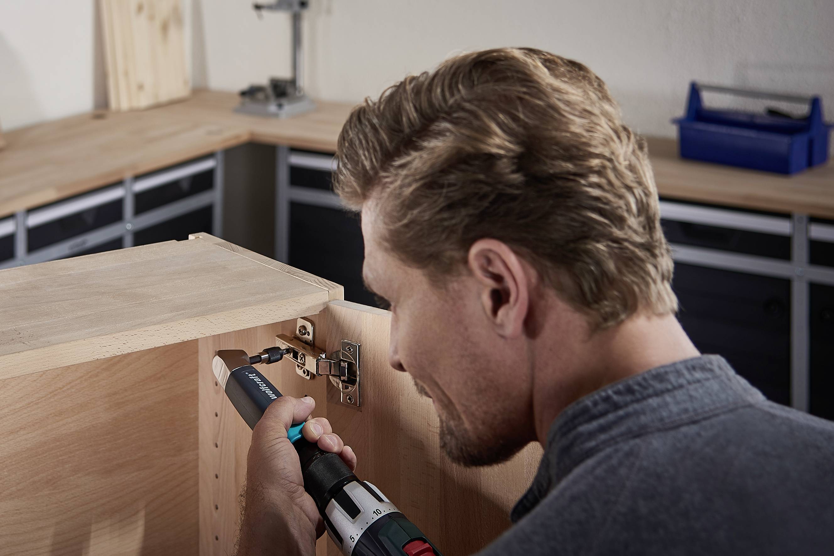 Ein Mann montiert ein Scharnier an einem hölzernen Schrankschrank mit einem Akkuschrauber. Werkzeuge und Werkbank im Hintergrund.