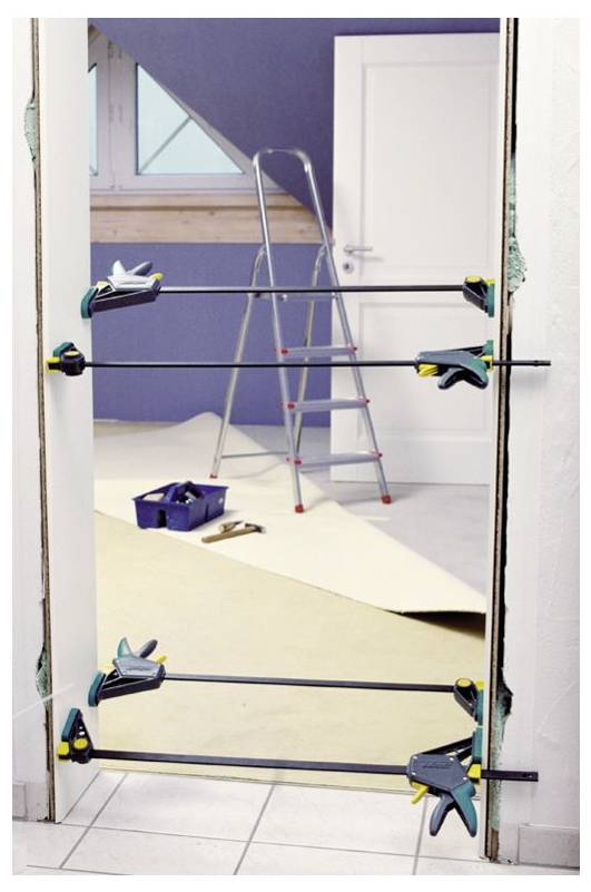 Ein Türrahmen, der während der Installation oder Reparatur mit Zwingen befestigt ist. Eine Trittleiter und eine Werkzeugkiste befinden sich im Hintergrund und deuten auf laufende Bauarbeiten hin.