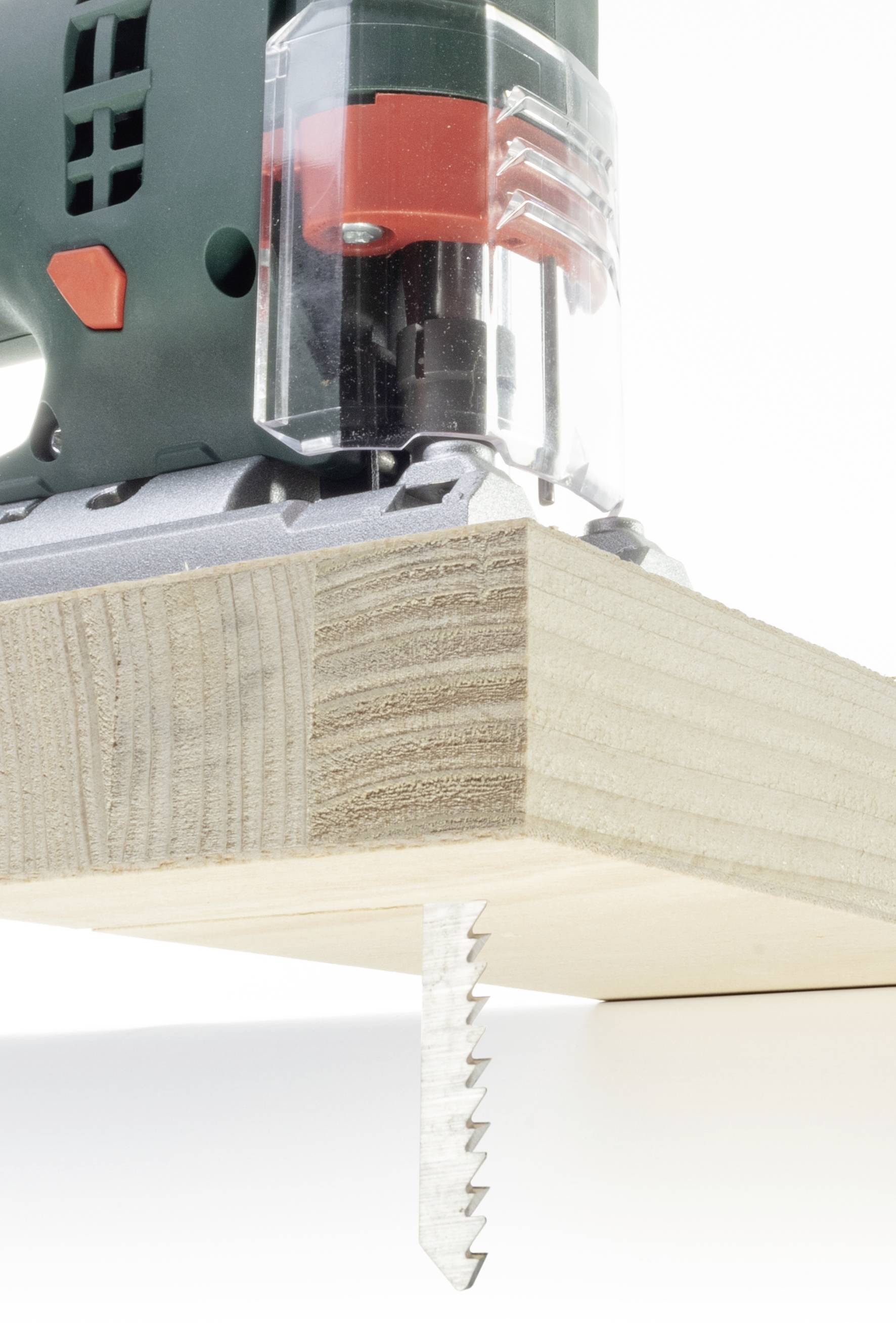 Eine Stichsäge schneidet durch ein Stück Holz, das hochkant positioniert ist. Die Säge hat ein grünes Gehäuse mit rotem Detail.