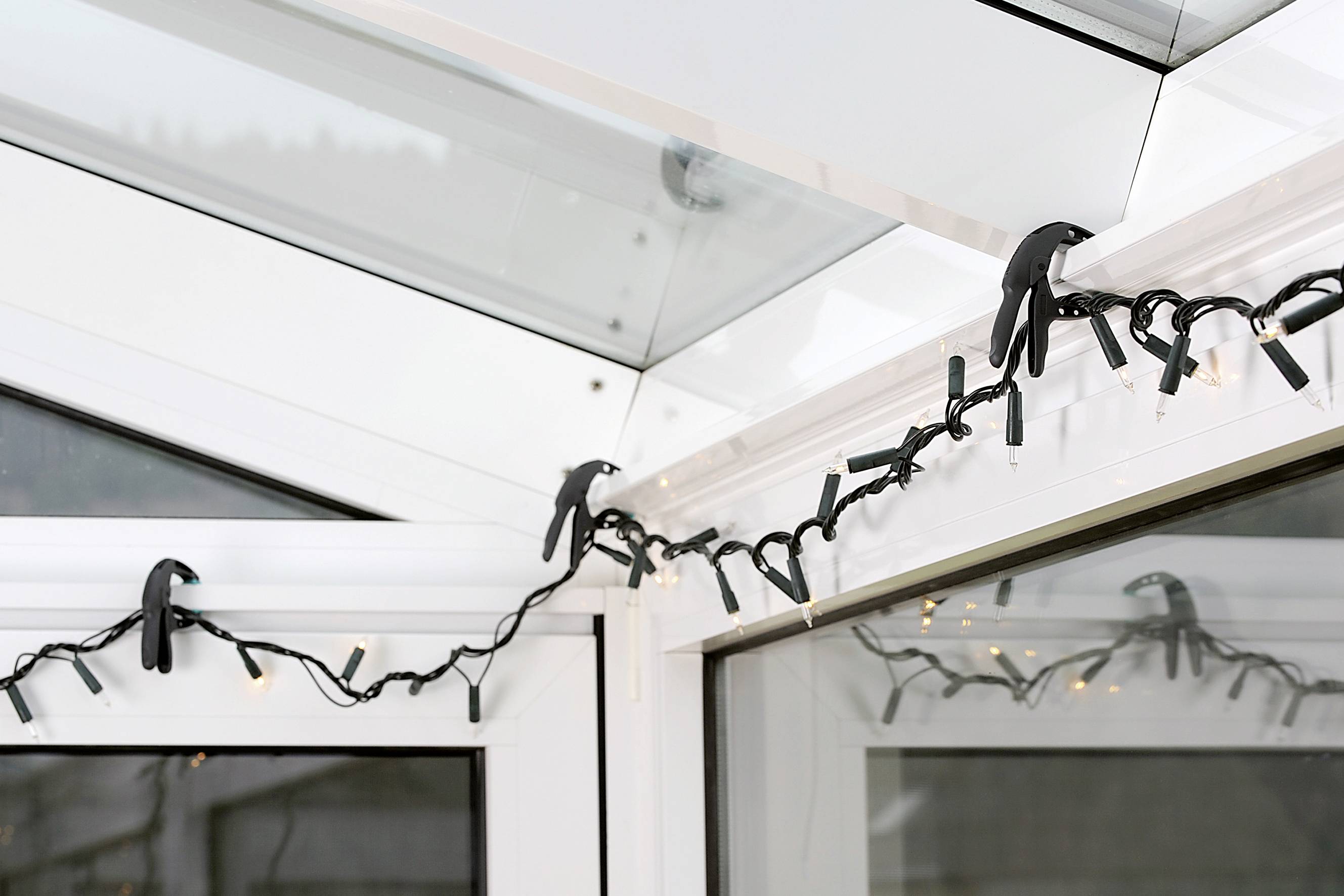Eine Lichterkette ist entlang der Ecke eines weißen Wintergartens angebracht, mit Clips an den Rahmen befestigt. Einige Lichter sind eingeschaltet.