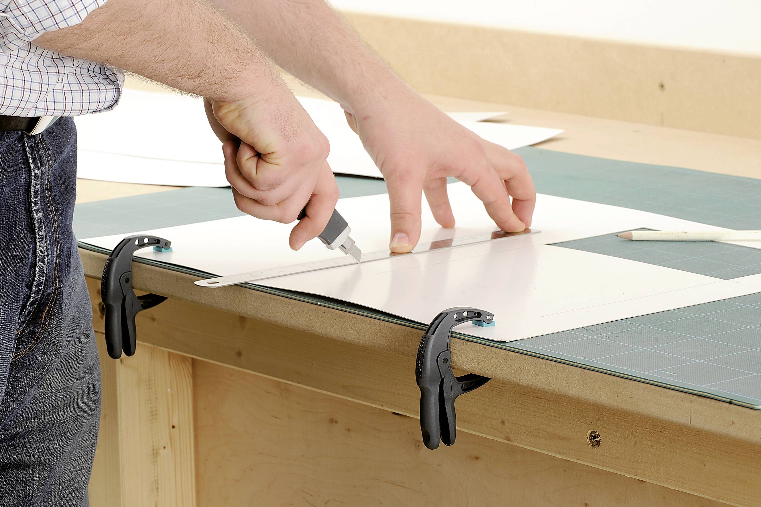 Eine Person schneidet mit einem Cutter-Messer präzise eine weiße Pappe, die auf einem Arbeitstisch mit Klemmen fixiert ist.