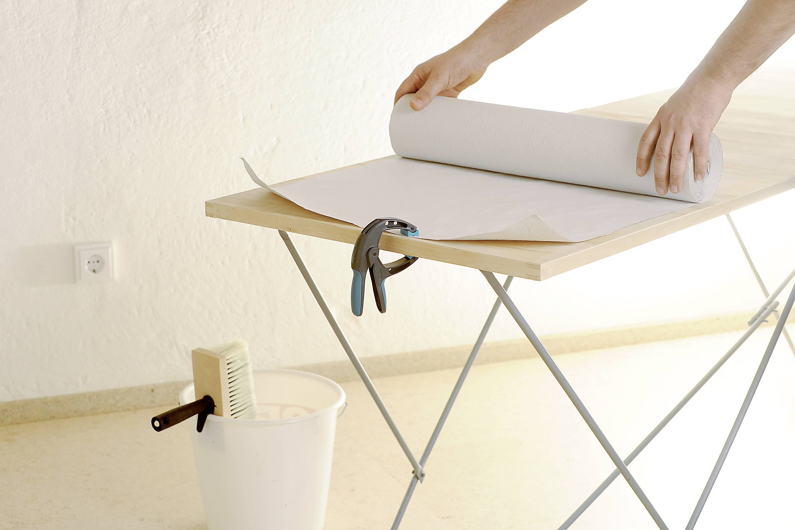 Eine Person rollt eine große Tapetenrolle auf einem Tisch ab, daneben steht ein Eimer mit Werkzeug.