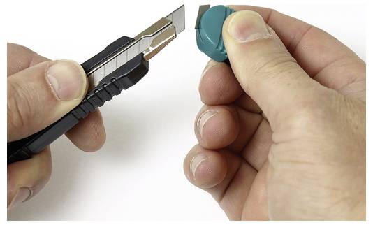 Eine Hand hält ein Cuttermesser und ist im Begriff, einen kleinen grünen Gegenstand, möglicherweise aus Plastik, zu schneiden, der von einer anderen Hand gehalten wird, was auf eine präzise Schneidaufgabe hindeutet.