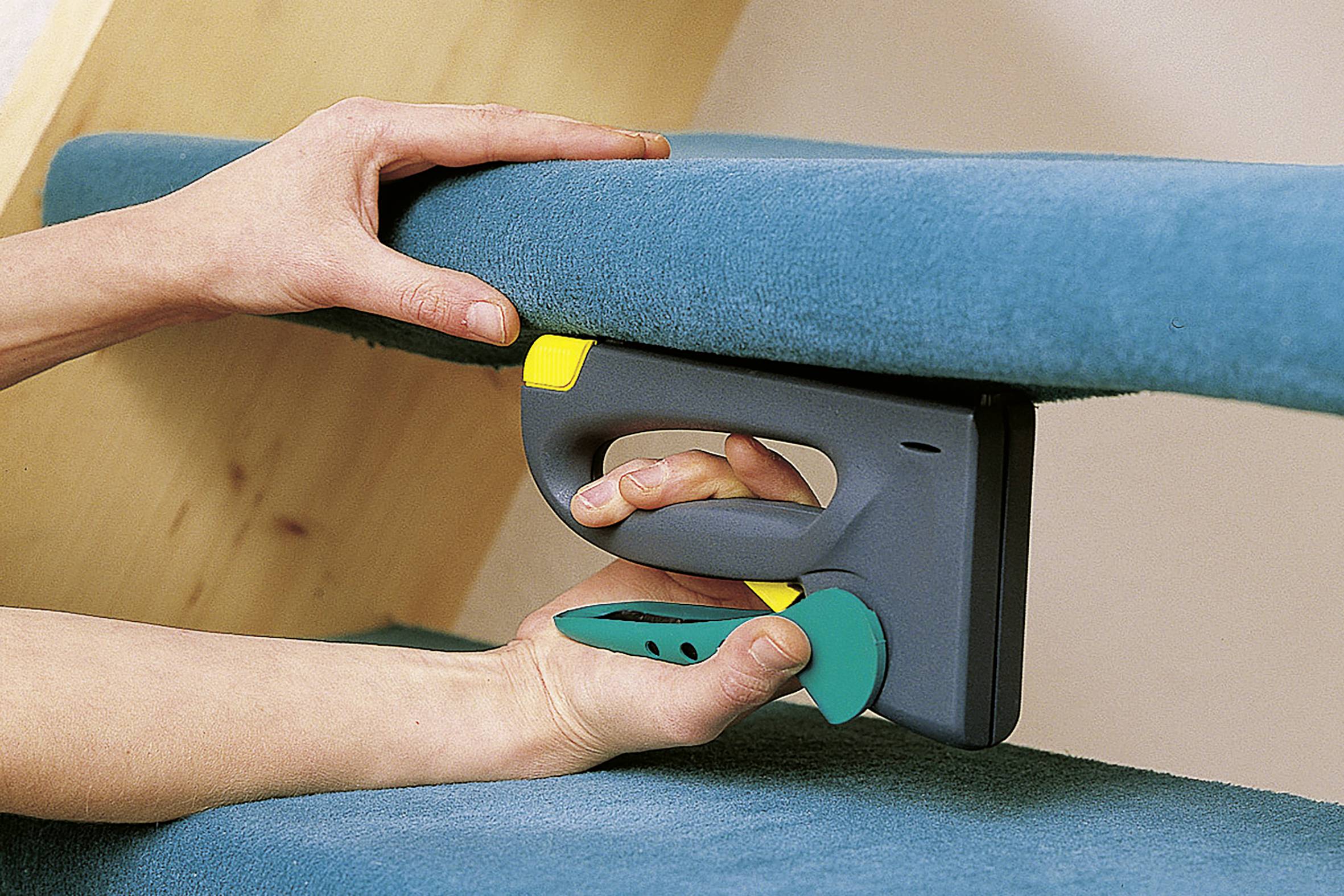 Person verwendet einen elektrischen Tacker, um blauen Stoff an einer Holzoberfläche zu befestigen. Hände halten den Stoff straff.