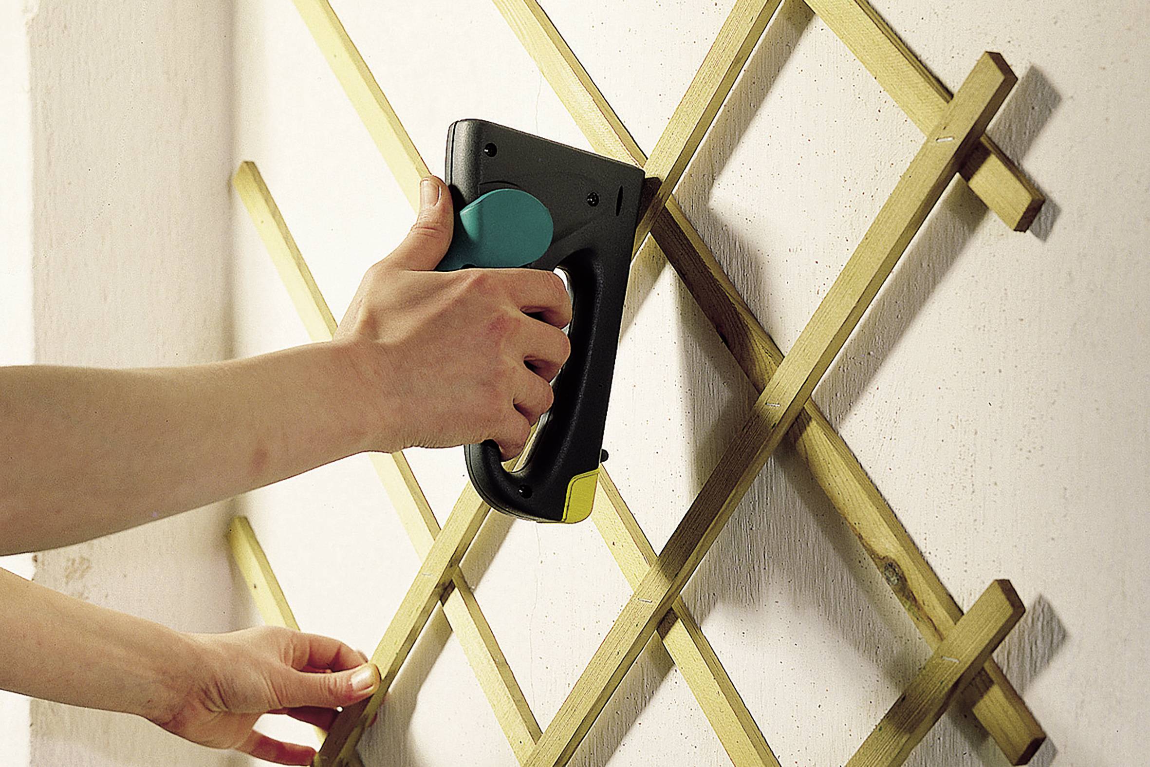 Eine Hand verwendet einen Tacker, um ein Holzgitter an einer Wand zu befestigen.