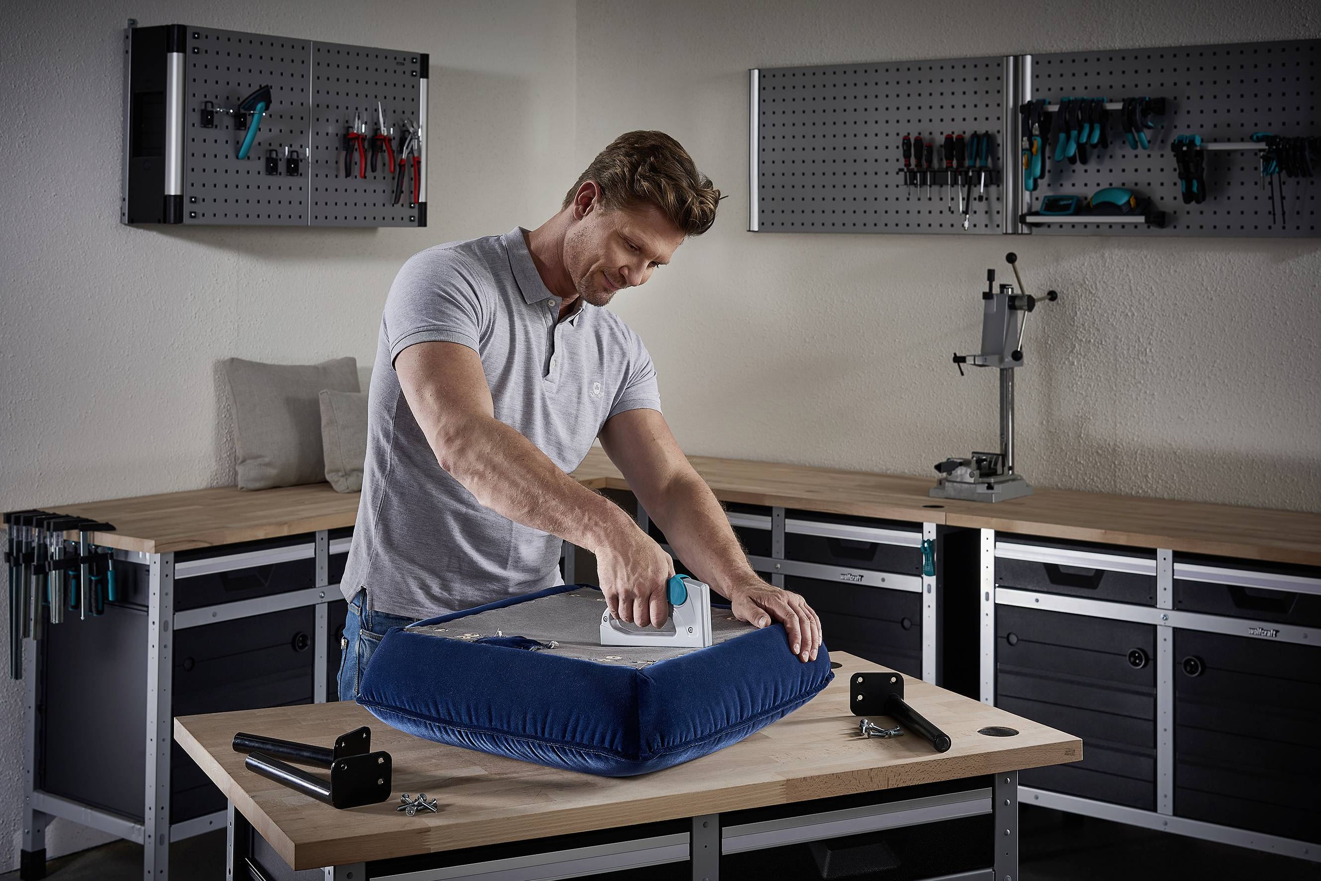 Ein Mann bezieht in einer Werkstatt einen Stuhl mit einem blauen Stoffbezug. Auf dem Tisch liegen Schrauben und Stuhlbeine bereit.