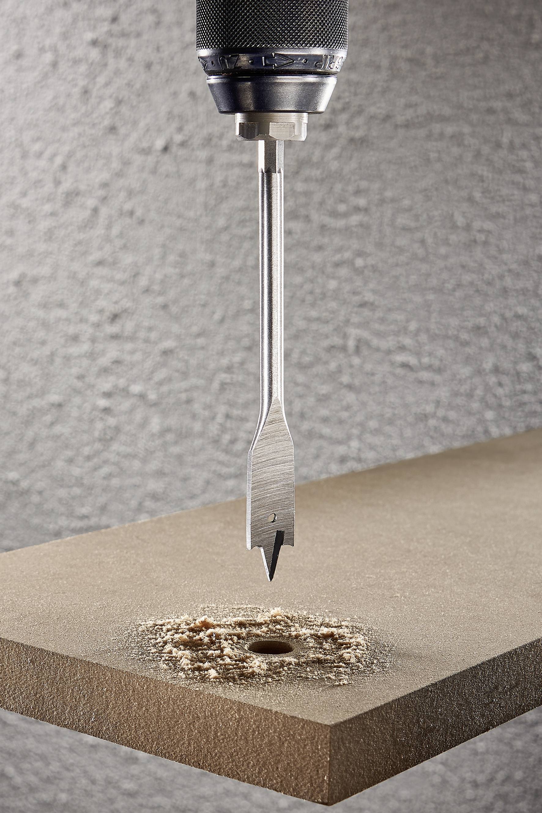 Eine Bohrmaschine mit flachem Bohrer bohrt ein Loch in eine Holzplatte, erzeugt Holzspäne auf einer grauen, strukturierten Oberfläche.