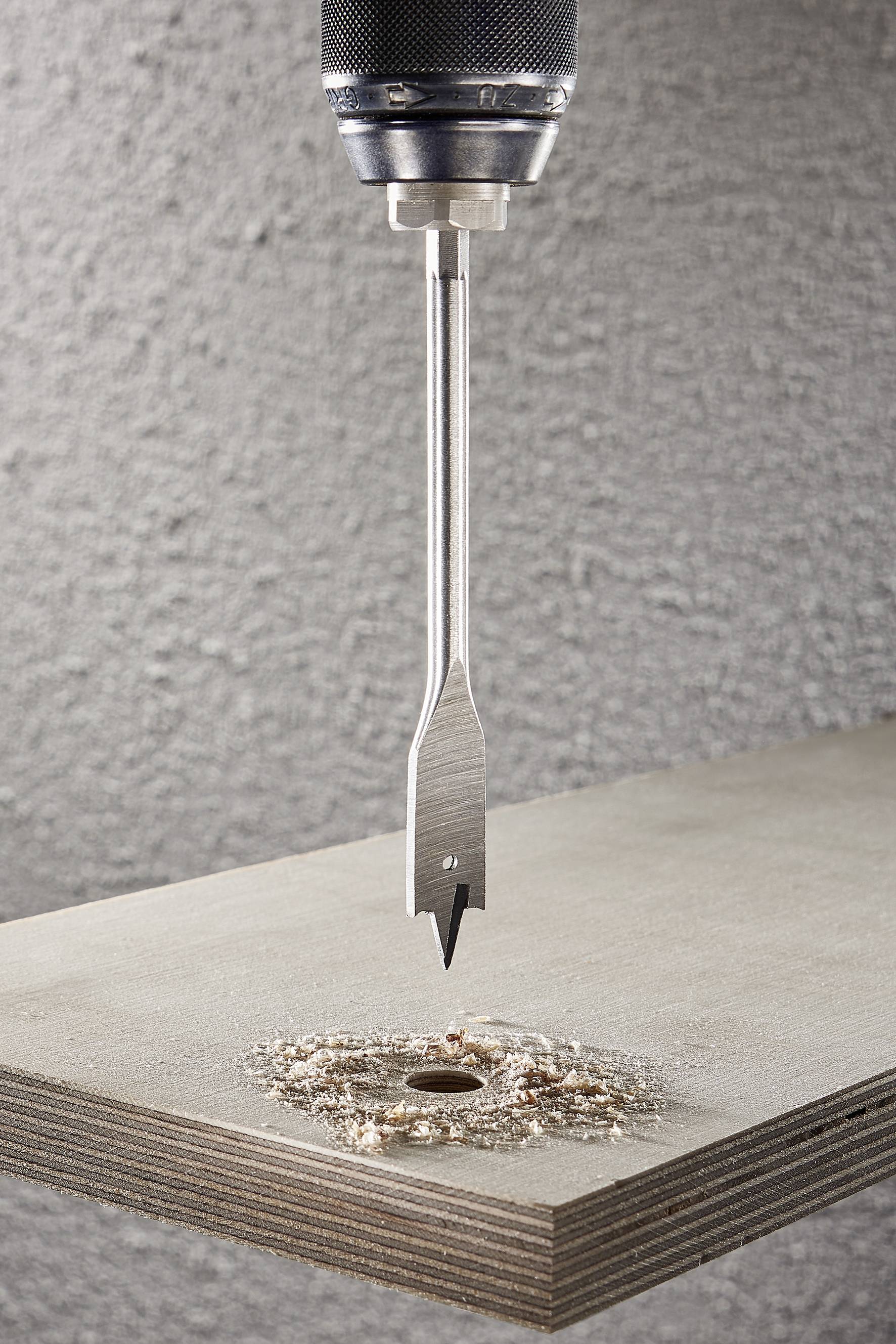 Ein Bohrer mit einem Flachbohrer bohrt ein Loch in ein Holzbrett. Holzspäne umgeben das Loch.