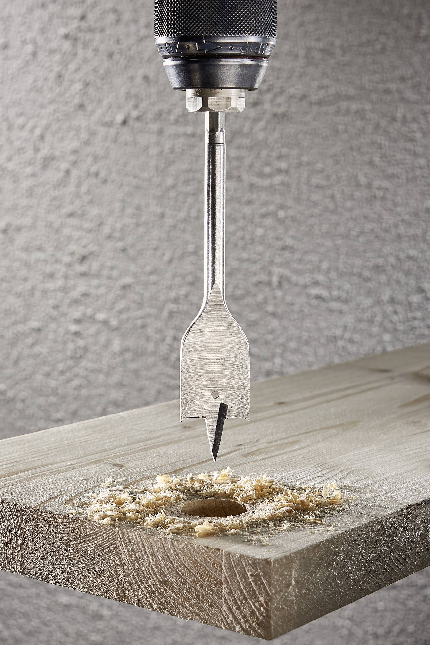 Ein Metallbohrer bearbeitet ein Holzbrett, wobei Holzspäne um das gebohrte Loch herum zu sehen sind.