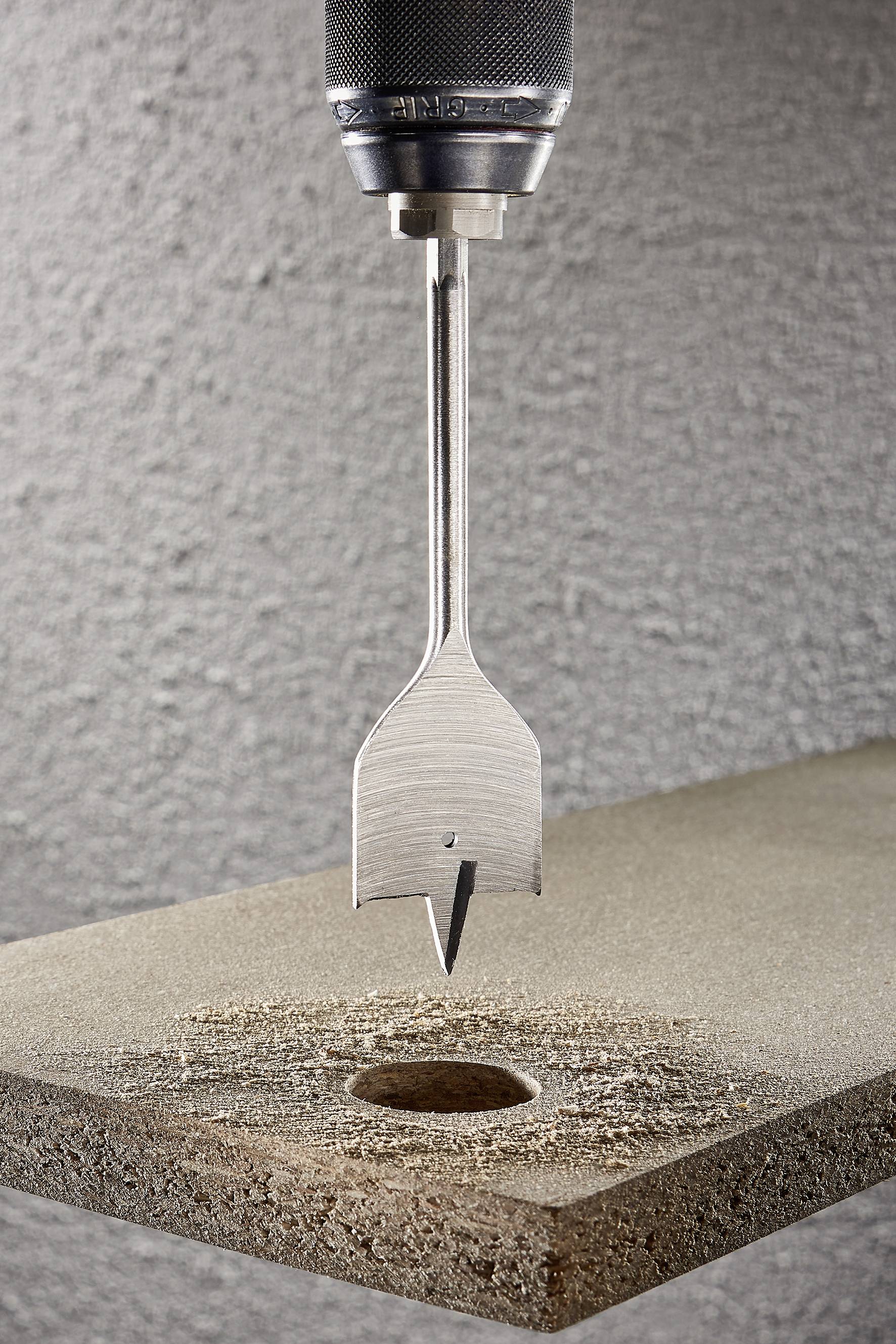 Bohrer mit Spatenbohrer durchbohrt Holzplatte vor grauer Wand. Der Bohrer ist mittig im Bild, zeigt Präzision und Handwerkskunst.