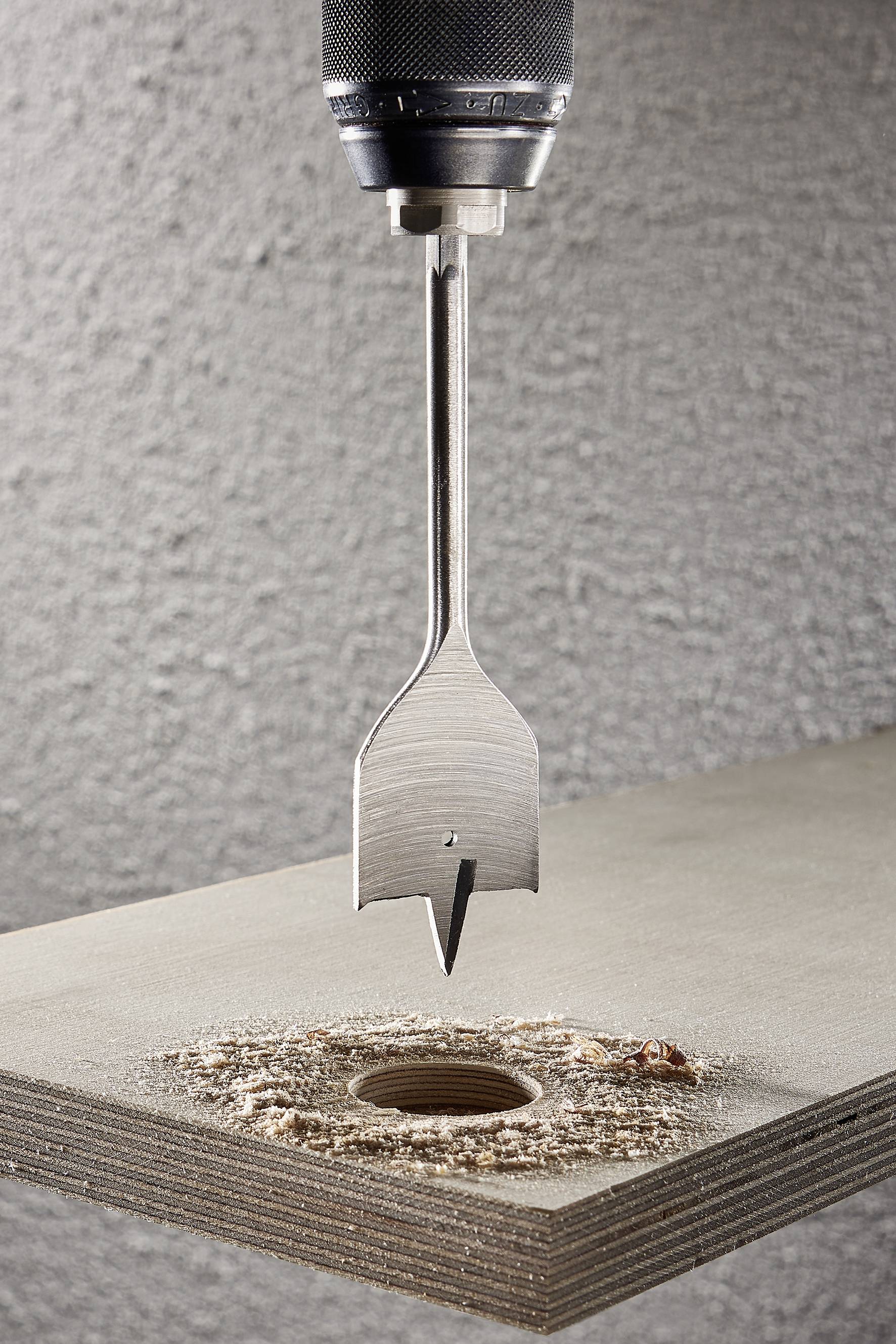 Ein Bohrer mit Flachfräsbohrer bohrt ein präzises Loch in ein Holzbrett. Holzspäne liegen um das Loch verstreut.