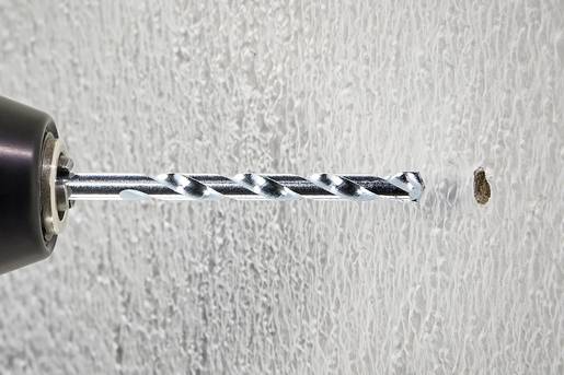 Ein Bohrer bohrt ein Loch in eine weiße, strukturierte Wand. Die Bohrspitze ist silbern und läuft sauber in die Wand ein.