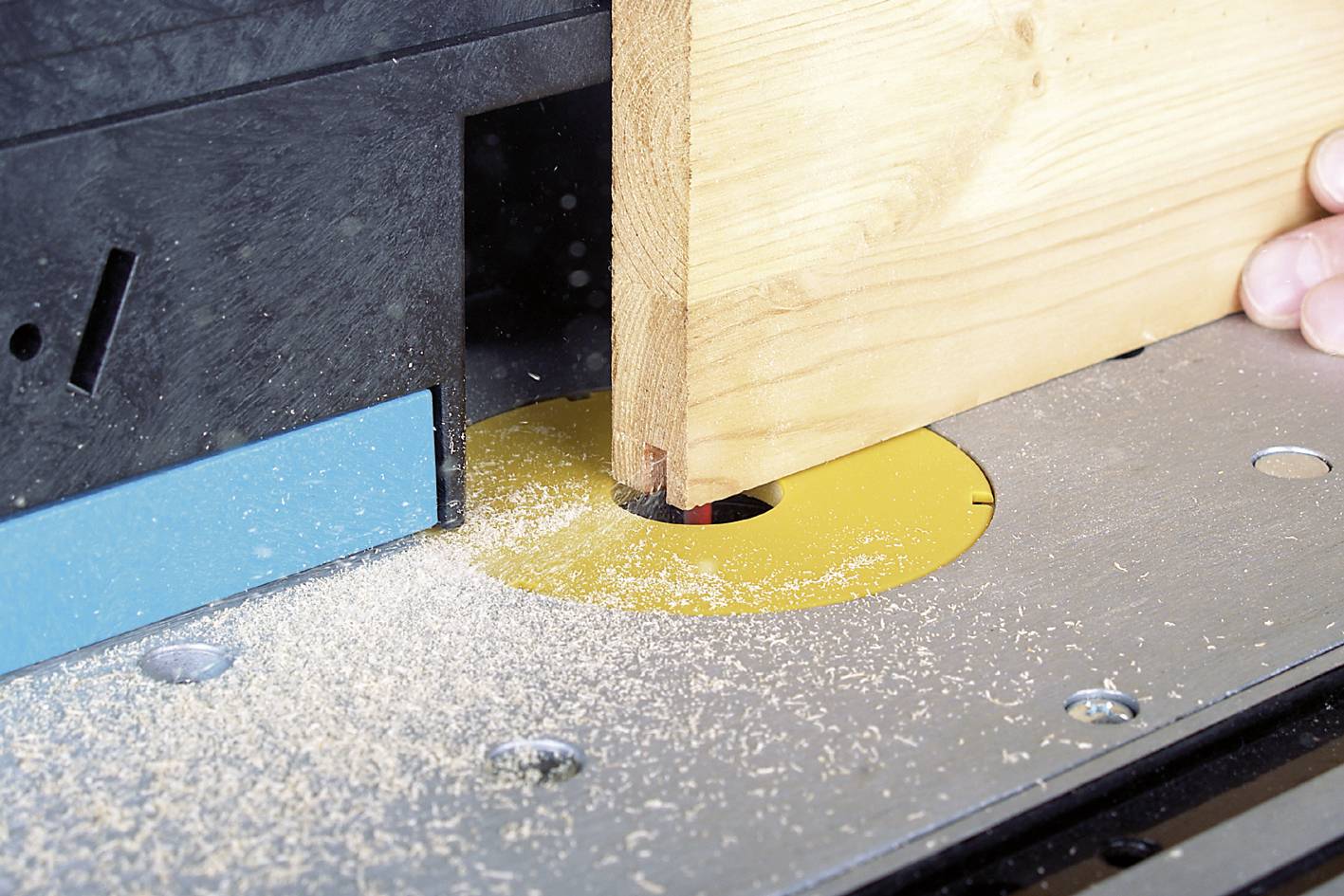 Eine Holzpaneele wird auf einem Tischfräser bearbeitet. Holzspäne sind um das Werkzeug verstreut. Eine Hand hält die Paneele.
