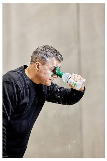 Eine Person in einem schwarzen Shirt benutzt eine Augenspülflasche, um ihre Augen zu reinigen, wobei sie ihren Kopf leicht nach vorne neigt, während Flüssigkeit herabfließt.
