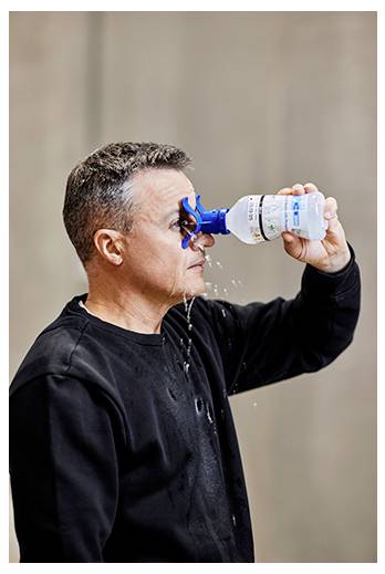 Eine Person benutzt eine Augenspülflasche und träufelt Flüssigkeit mit der Ausgießspitze eines kleinen Behälters in ihr Auge, während die Flüssigkeit an ihrem Gesicht herabläuft.