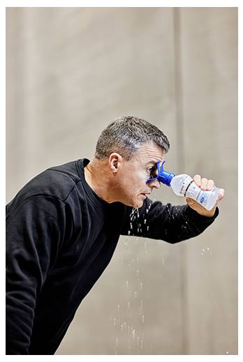 Ein Mann in einem schwarzen Oberteil benutzt eine Augenspülflasche, um sein Auge zu spülen. Wasser rinnt von der Flasche herunter und fließt über sein Gesicht und seine Kleidung.