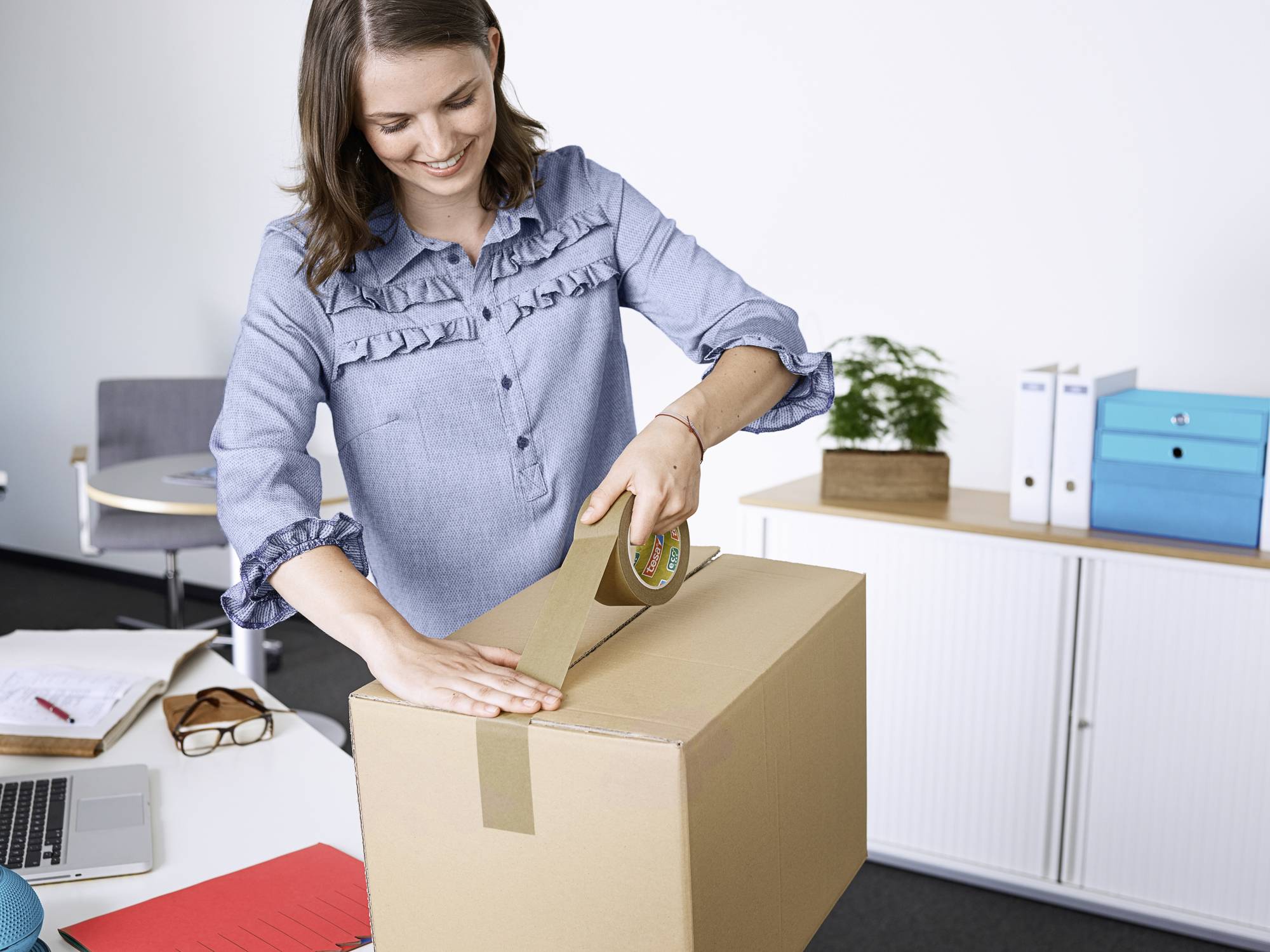 Eine Frau klebt einen Karton mit Klebeband zu. Sie ist in einem Büro mit Schreibtisch, Laptop, Büchern und Pflanzen im Hintergrund.