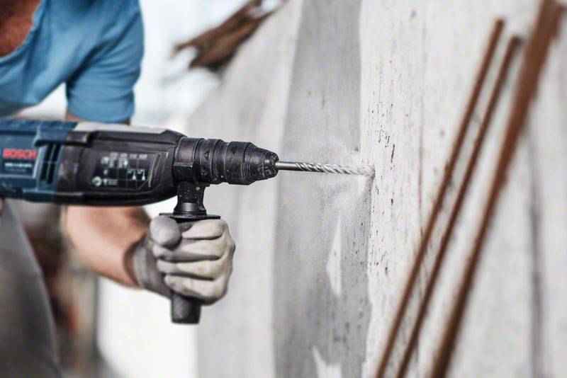 Mann im blauen Shirt bohrt mit einer Elektromaschine ein Loch in eine Betonwand. Schutzbekleidung, einschließlich Handschuhe, werden getragen.