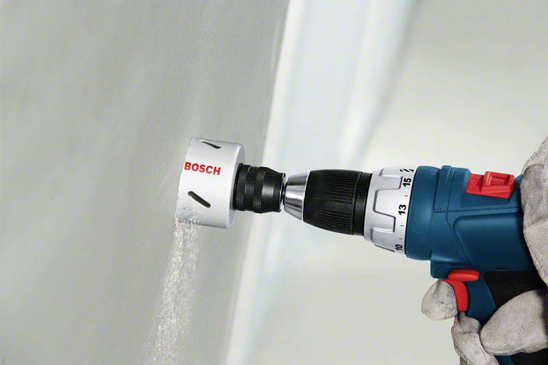 Bohrmaschine mit Lochsägeaufsatz bohrt in eine Wand, markiert mit 'Bosch'. Eine Hand mit Handschuh hält die Maschine.