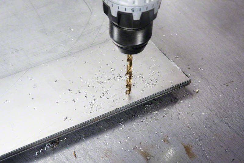 Eine Bohrmaschine bohrt ein Loch in ein Metallblech auf einer werkstattähnlichen Oberfläche und erzeugt Metallspäne.