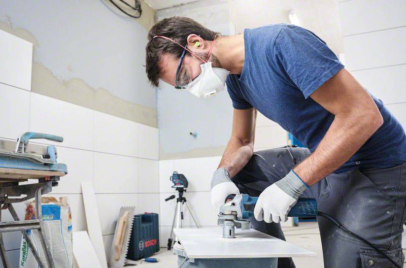 Ein Handwerker trägt Schutzbrille und Maske, während er mit einer Säge an einer Fliese arbeitet. Im Hintergrund sind Werkzeuge sichtbar.
