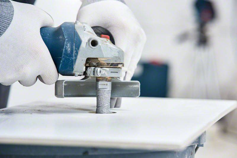 Eine Person bearbeitet eine weiße Fliese mit einem Elektrowerkzeug, trägt weiße Schutzhandschuhe und arbeitet in einer Werkstattumgebung.