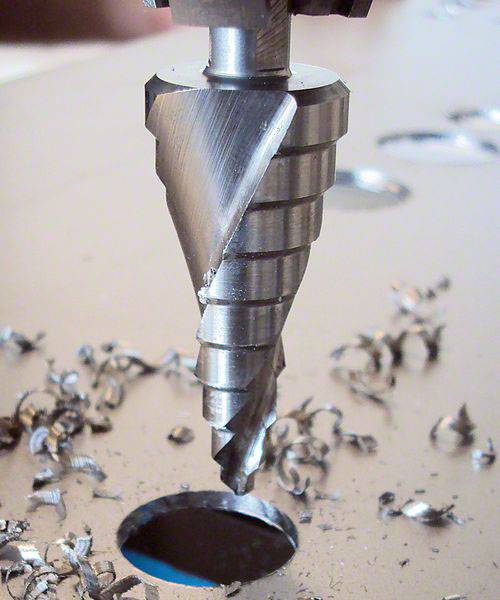 Ein Stufenbohrer durchbohrt eine Metallplatte. Metallspäne sind um das Loch verteilt.