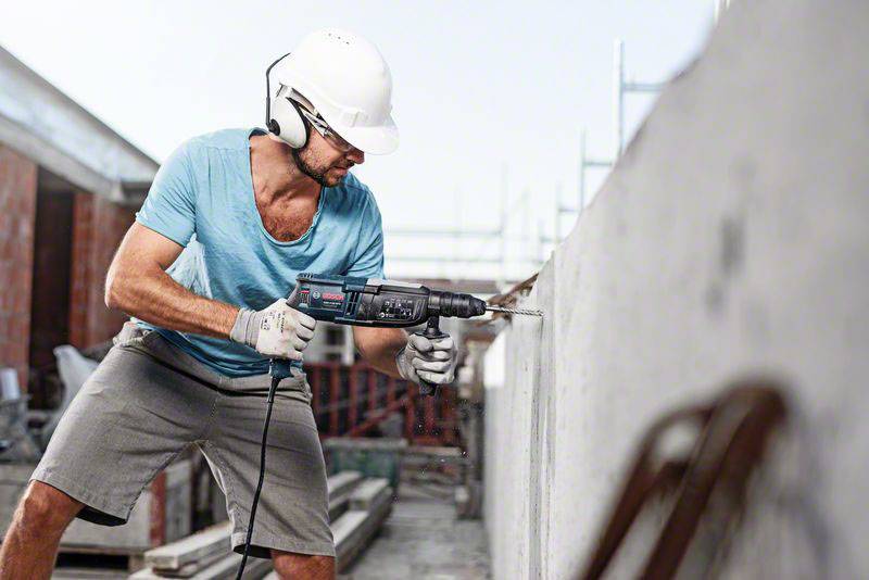 Ein Bauarbeiter mit Helm und Ohrenschützern bohrt mit einer Elektromaschine ein Loch in eine Betonwand auf einer Baustelle.