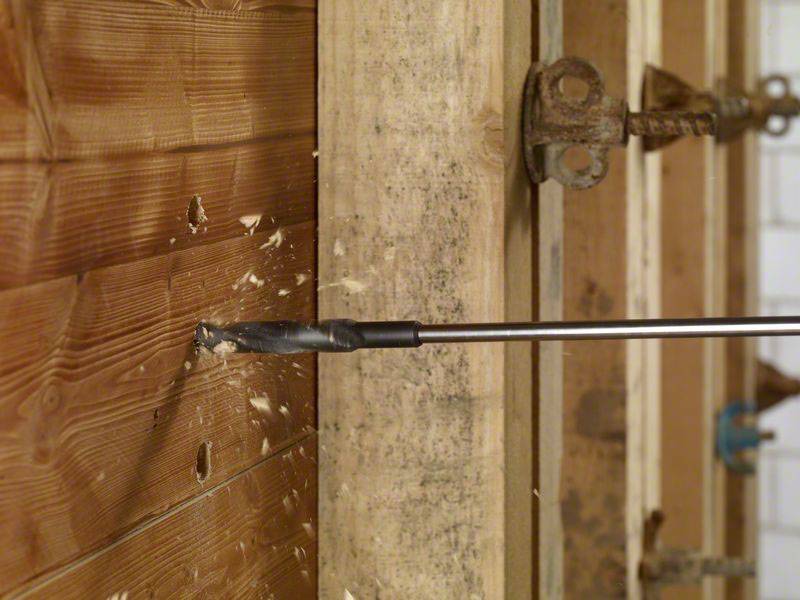 Ein Bohrer durchbohrt eine Holzwand, dabei fliegen Holzspäne. Im Hintergrund sind Teile einer strukturellen Holzkonstruktion sichtbar.