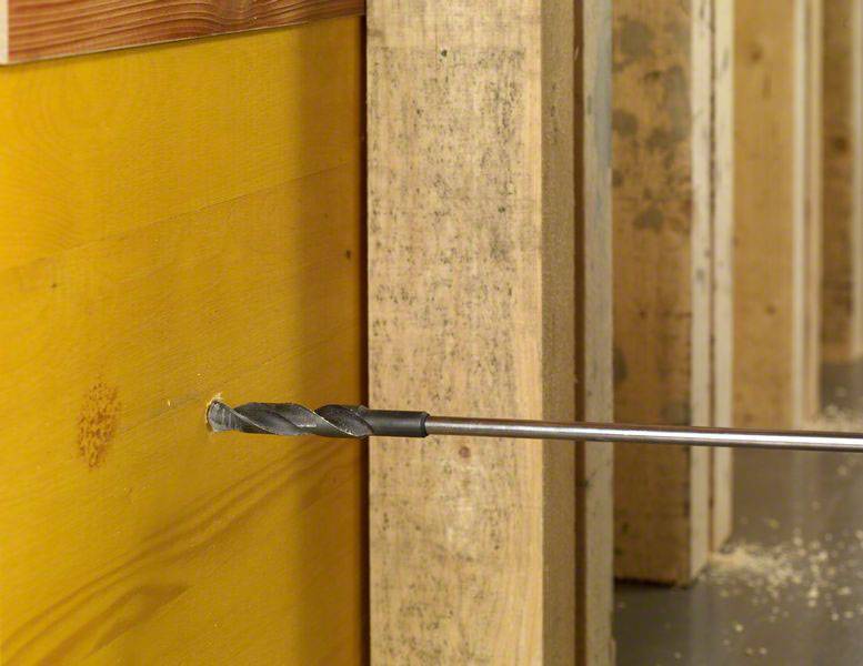 Ein Bohrer durchbohrt ein gelbes Brett aus Holz, umrahmt von Holzbalken, in einer Werkstattumgebung.