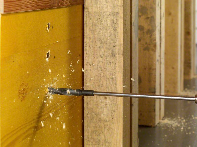 Ein Bohrer durchbohrt ein Holzbrett. Holzspäne fliegen heraus. Im Hintergrund sind weitere Bretter zu sehen.