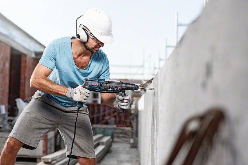 Ein Bauarbeiter mit Schutzhelm und Kopfhörern bohrt mit einer Bohrmaschine in eine Betonwand auf einer Baustelle.