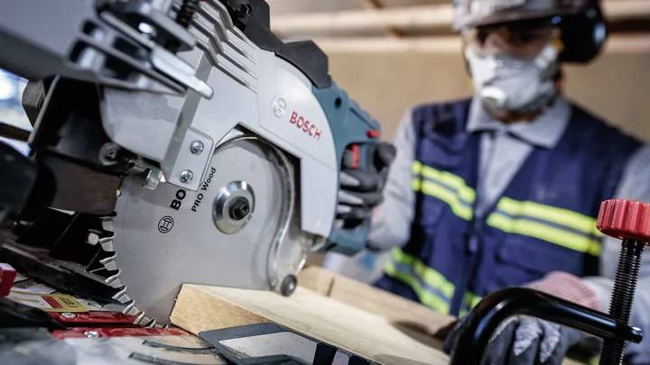 Ein Arbeiter sägt Holz mit einer Bosch-Kreissäge. Der Arbeiter trägt Schutzkleidung einschließlich Helm, Schutzbrille und Maske.