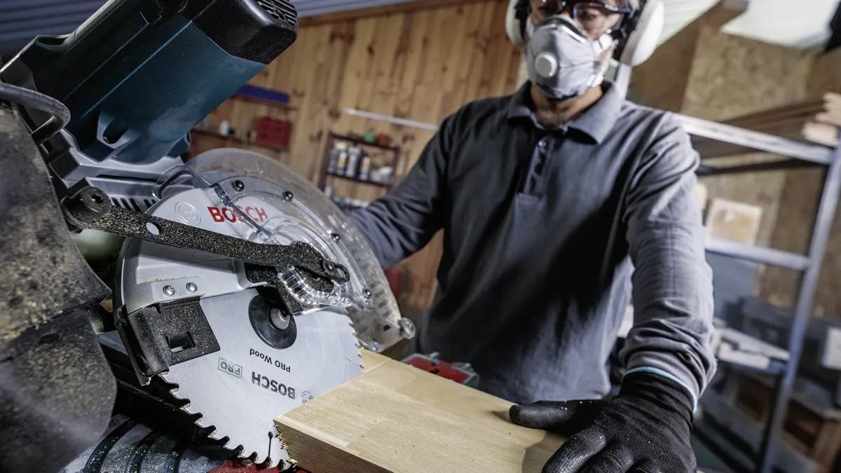 Eine Person mit Sicherheitsausrüstung bedient eine Kreissäge, um ein Holzbrett in einer Werkstattumgebung zu schneiden.