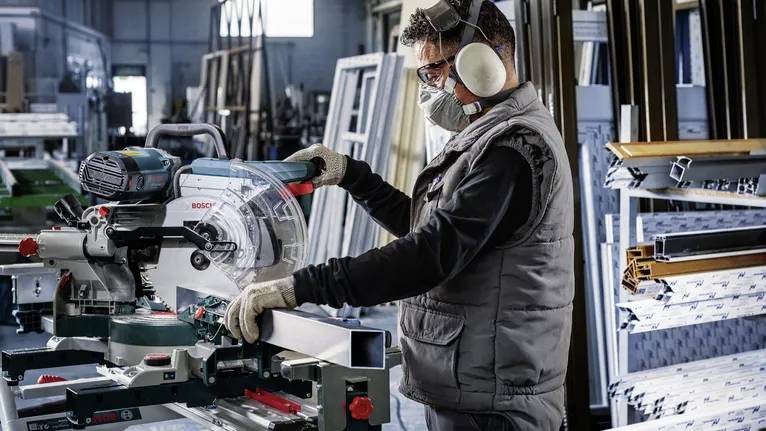 Eine Person mit Schutzausrüstung bedient eine Kreissäge in einer Werkstatt mit Metallrahmen und Werkzeugen im Hintergrund.