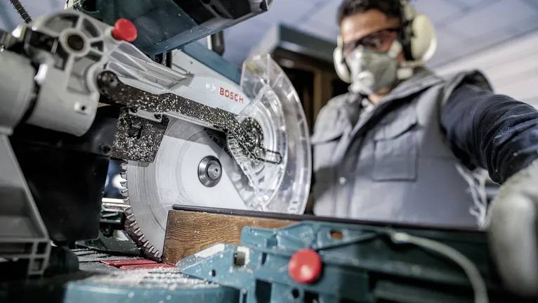 Ein Arbeiter bedient eine Kreissäge und schneidet ein Stück Holz. Der Arbeiter trägt Schutzausrüstung, einschließlich Gehörschutz und einer Maske.