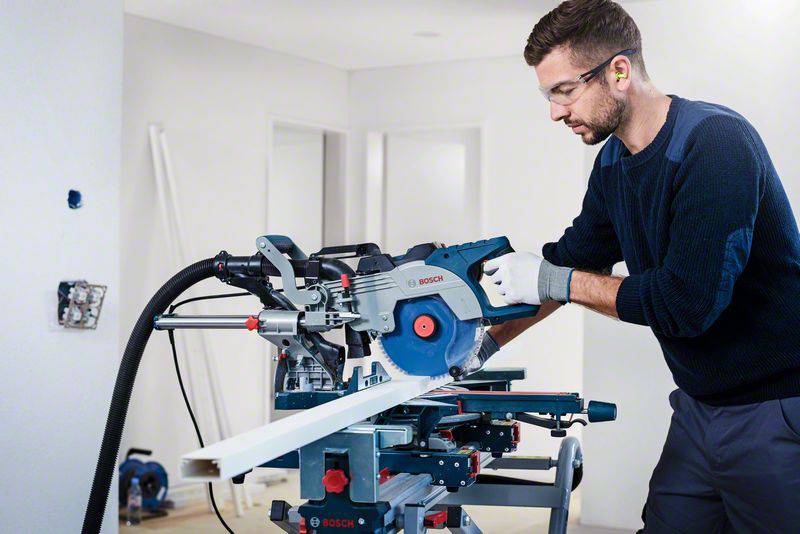 Ein Mann bedient in einem hellen Raum eine elektrische Säge, um ein weißes Holzbrett zu schneiden. Er trägt Schutzbrille und Gehörschutz.