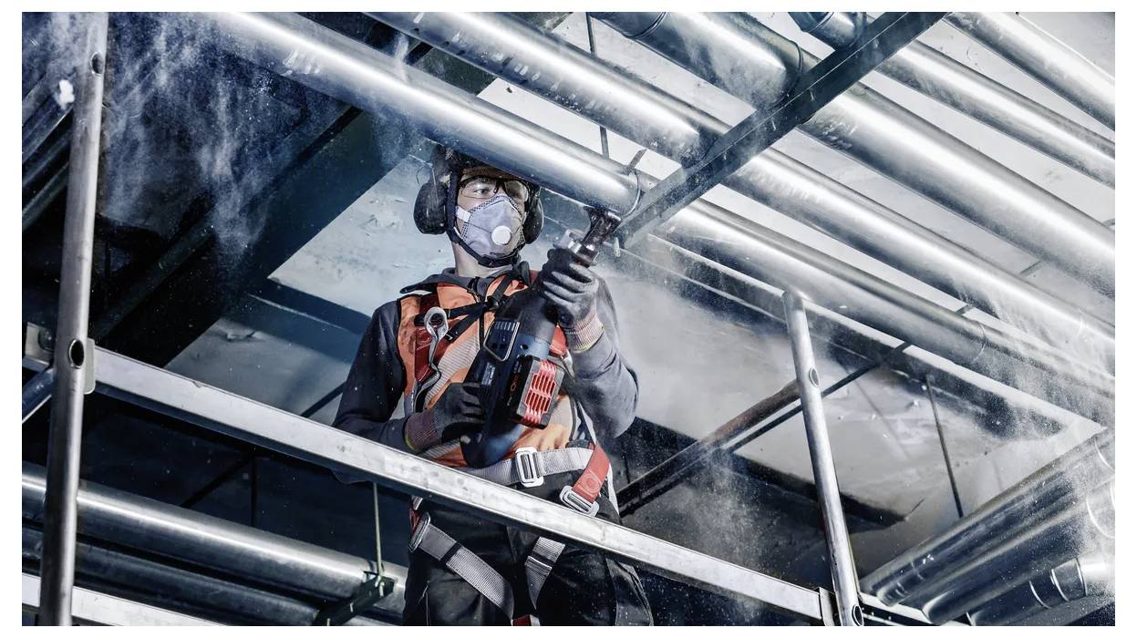 Arbeiter in Schutzausrüstung nutzt einen Hochdruckschlauch auf industriellem Gerüst, umgeben von Metallrohren. Sicherheitsausrüstung ist sichtbar.