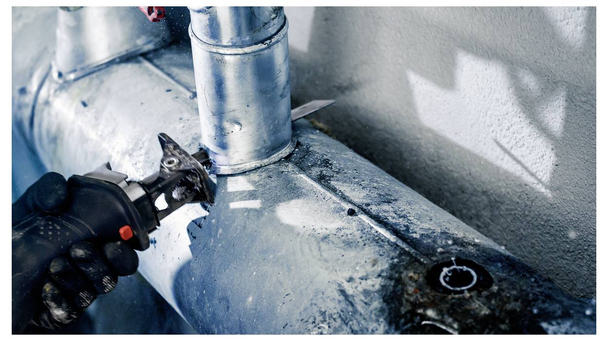 Eine Person verwendet ein Werkzeug mit elektrischem Antrieb, um ein Metallrohr zu schneiden, wobei Funken sichtbar sind. Die Szene deutet auf industrielle Arbeiten oder laufende Installationsarbeiten hin.