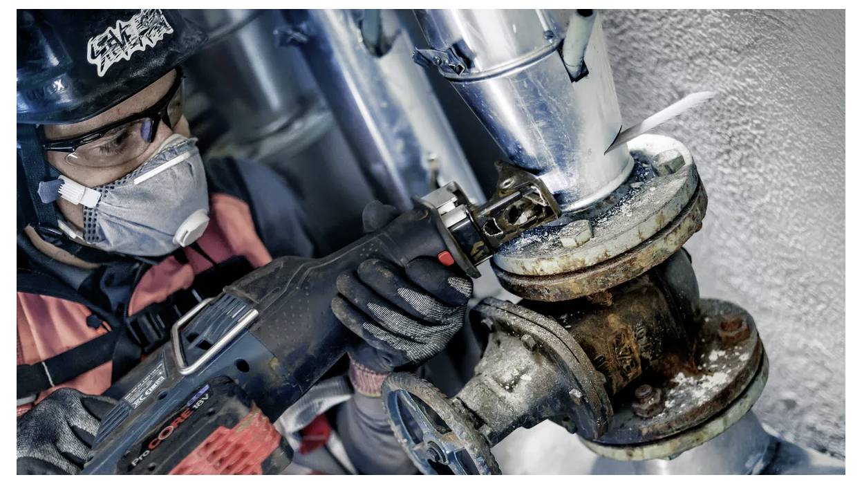 Ein Arbeiter in Sicherheitsausrüstung, einschließlich Helm und Maske, verwendet ein Elektrowerkzeug, um ein verrostetes Metallrohr in einer industriellen Umgebung zu durchtrennen.