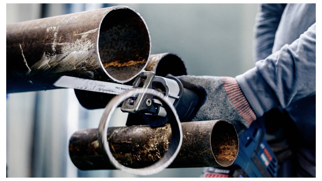 Eine Person in Schutzhandschuhen verwendet eine Handsäge, um eine große, verrostete Metallröhre präzise durchzuschneiden und konzentriert sich dabei auf einen exakten Schnitt.