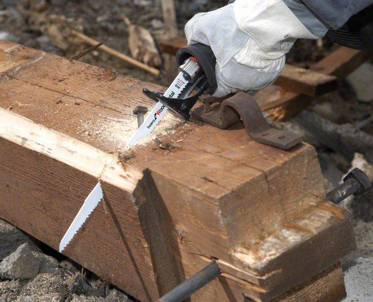 Eine Person sägt mit einer elektrischen Säge ein dickes Holzbrett. Späne fliegen, und die Umgebung wirkt industriell.