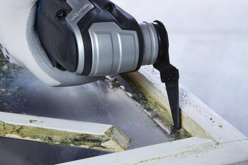 Eine Hand arbeitet mit einem elektrischen Gerät, um einen Fensterrahmen zu reparieren. Das Werkzeug entfernt beschädigte Holzteile.