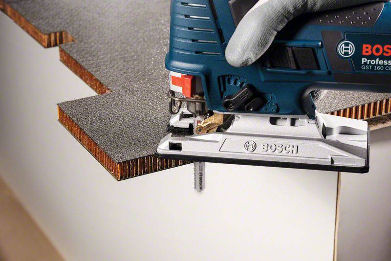 Eine elektrische Stichsäge mit einer blauen Gehäuse schneidet eine Holzplatte. Eine behandschuhte Hand hält die Säge fest.