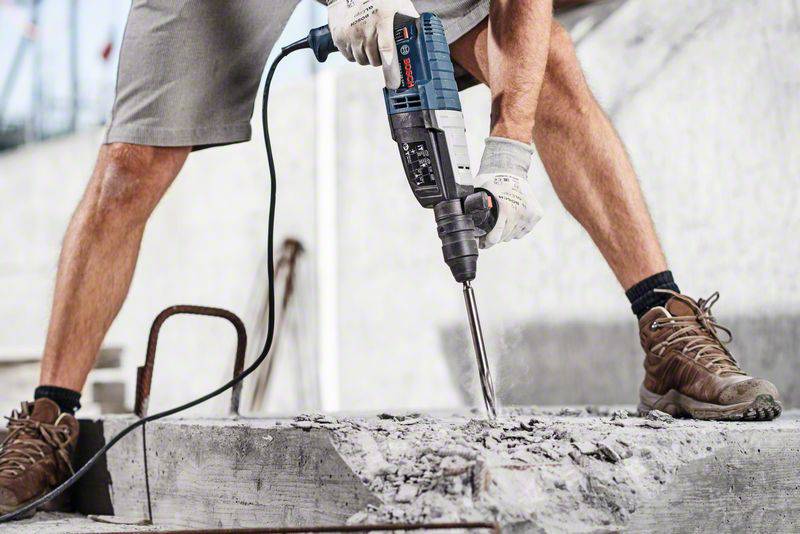 Eine Person in Arbeitshandschuhen benutzt einen Bohrhammer, um Beton zu bearbeiten. Im Hintergrund sind Baugerüste zu sehen.
