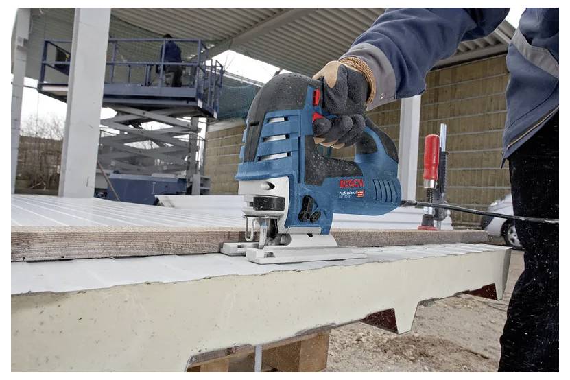 Eine Person mit Handschuhen verwendet eine Stichsäge, um ein Holzbrett im Freien zu schneiden. Ein Hubarbeitsbühne und ein weiterer Arbeiter sind im Hintergrund sichtbar.