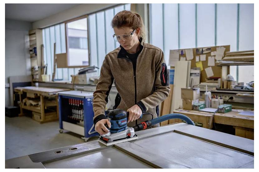 Ein Arbeiter in einer Schutzbrille verwendet einen tragbaren Schleiffer auf einem großen Holzpaneel in einer Werkstatt.
