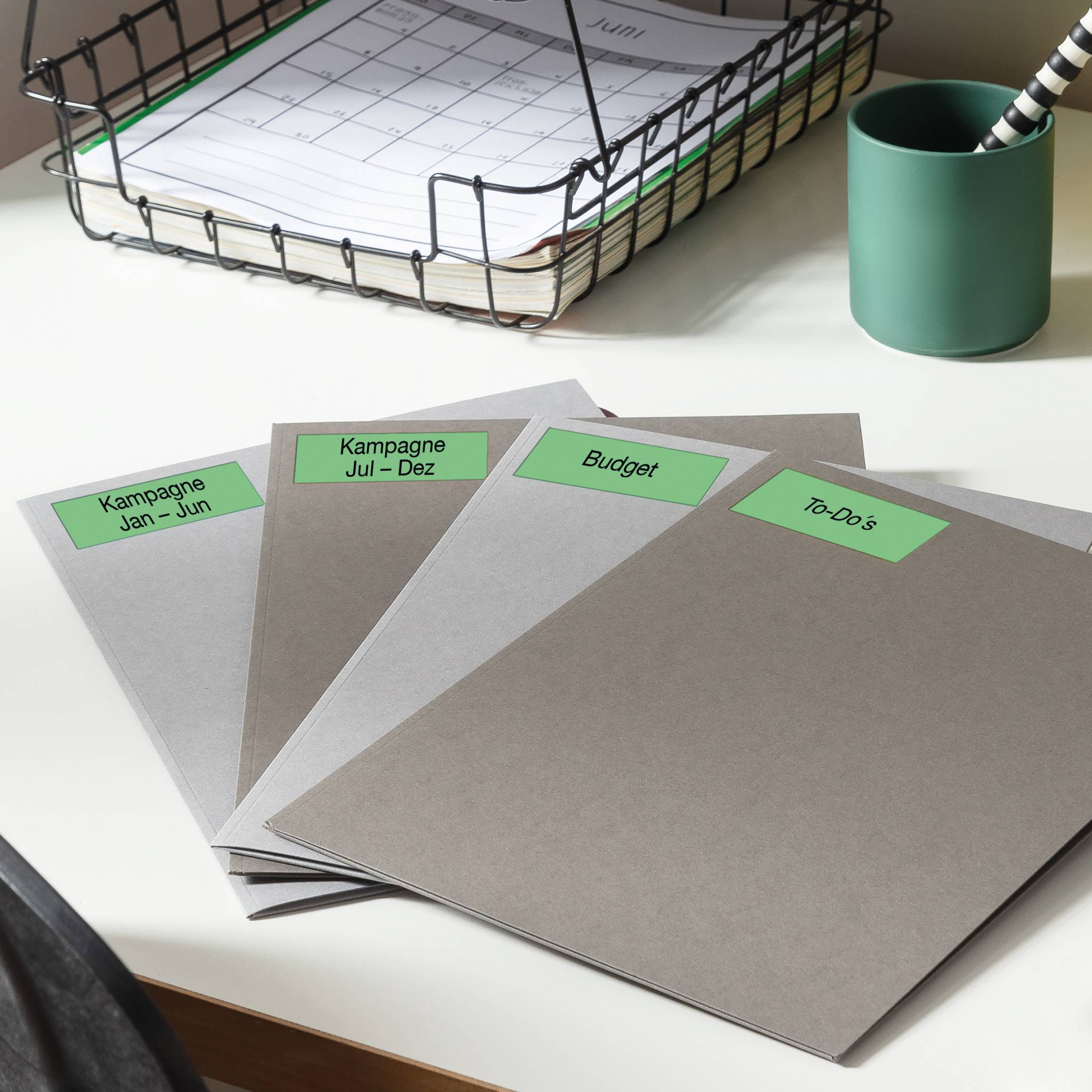 Vier Mappen auf einem Schreibtisch, beschriftet mit 'Kampagne Jan - Jun', 'Kampagne Jul - Dez', 'Budget', 'To Do's'. Im Hintergrund ein Kalender.