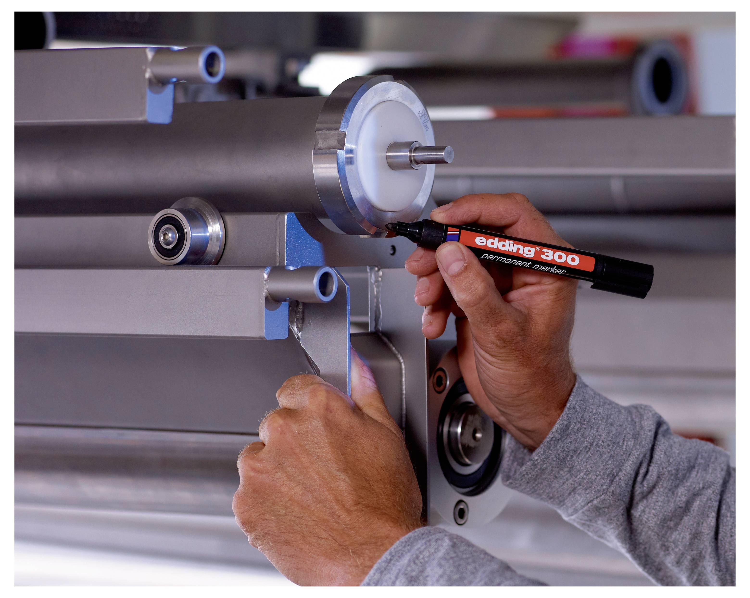 Ein Arbeiter markiert ein metallisches Maschinenteil mit einem schwarzen „edding 300"-Marker, während er das Bauteil an Ort und Stelle hält.