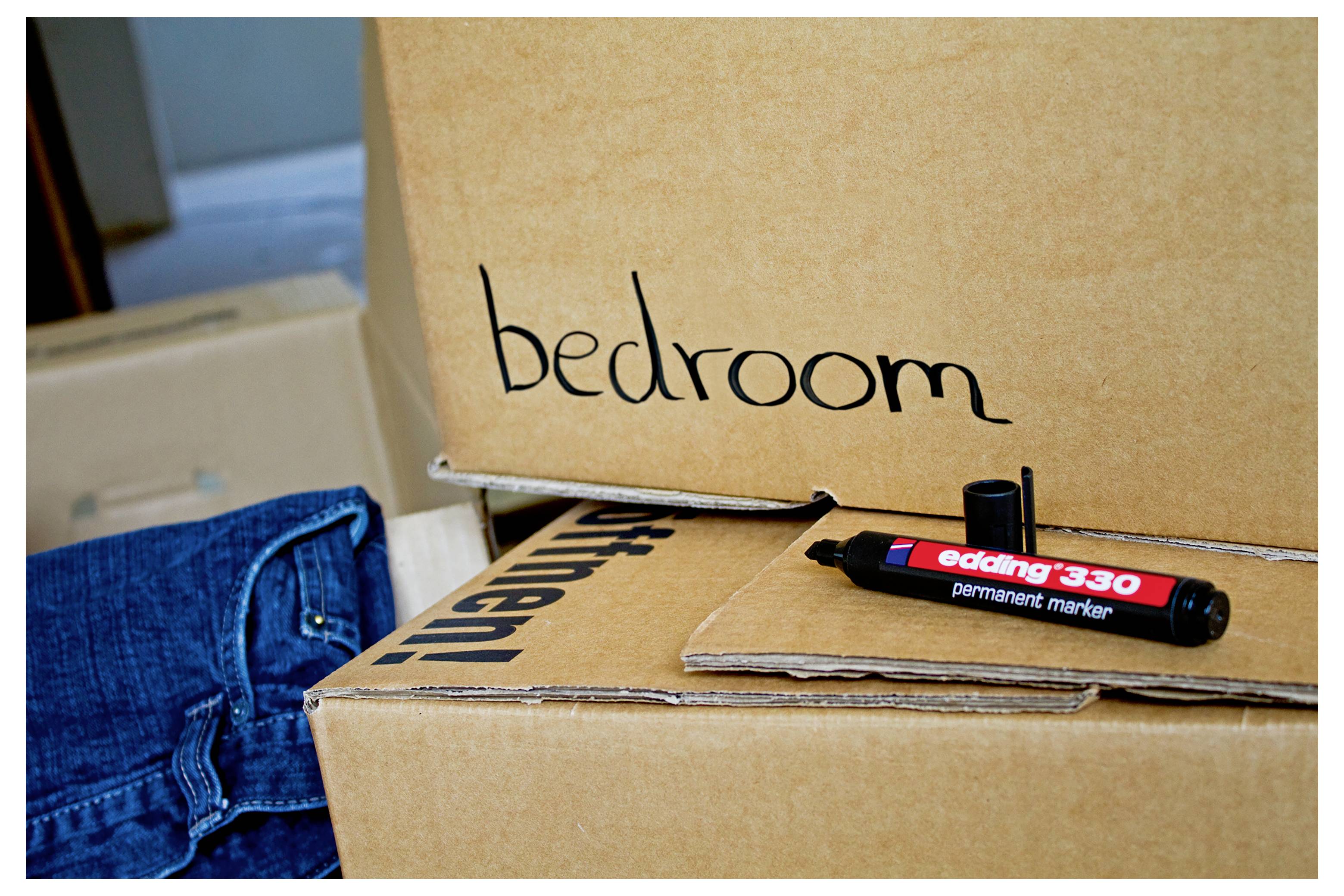 Umzugskartons mit der Beschriftung "Schlafzimmer" und "Küche", ein permanenter Marker in der Nähe und zusammengefaltete Jeans, was auf das Packen für einen Umzug hindeutet.